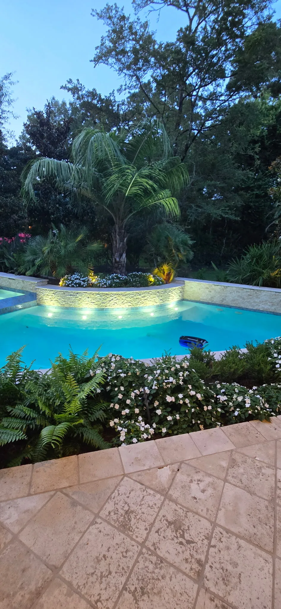 Backyard pool at dusk with LED lighting glowing along a stone water feature wall